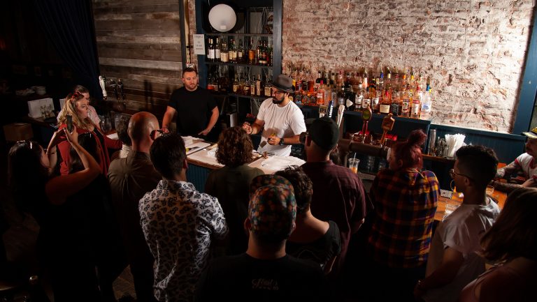 A group gathered around a bar, featuring an exposed brick wall, watch 2 men behind the bar give instruction at a Bar Methods summit in NYC