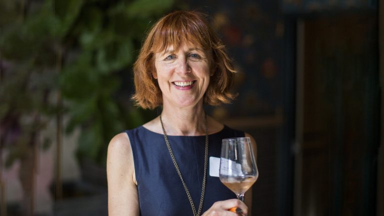 Mary Gormam-McAdams faces the camera with a glass of white wine held in front of her. She has a wide smile and a navy blue sleeveless blouse on.