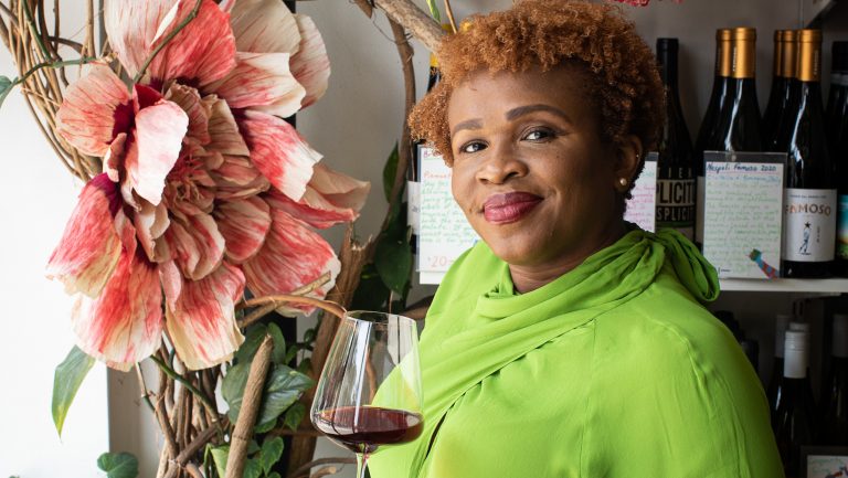 Deniece Bourne offers a gentle half-smile to the camera while holding a glass of read wine and wearing a vibrant lime green sop. There is a large blooming flower over her right shoulder.
