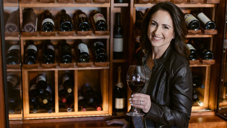 Julie Cohen theobald is wearing a black leather jacket and is holding a glass of red wine. She stands in front of filled wooden racks of wine bottles.