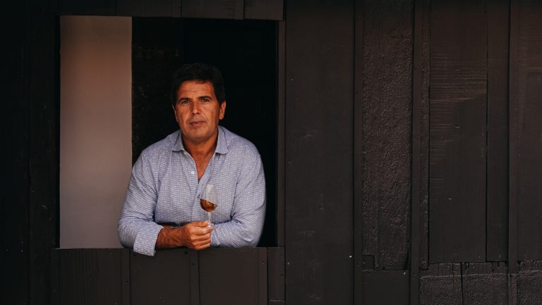 Francisco Albuquerque sits against a dark background wearing a light blue oxford shirt and holds a glass of red wine.
