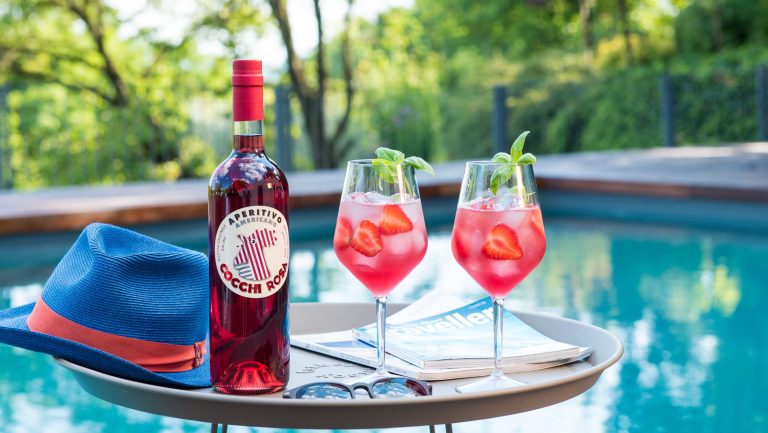 A poolside pictures of a bottle of Cocchi Rosa next to 2 cocktails