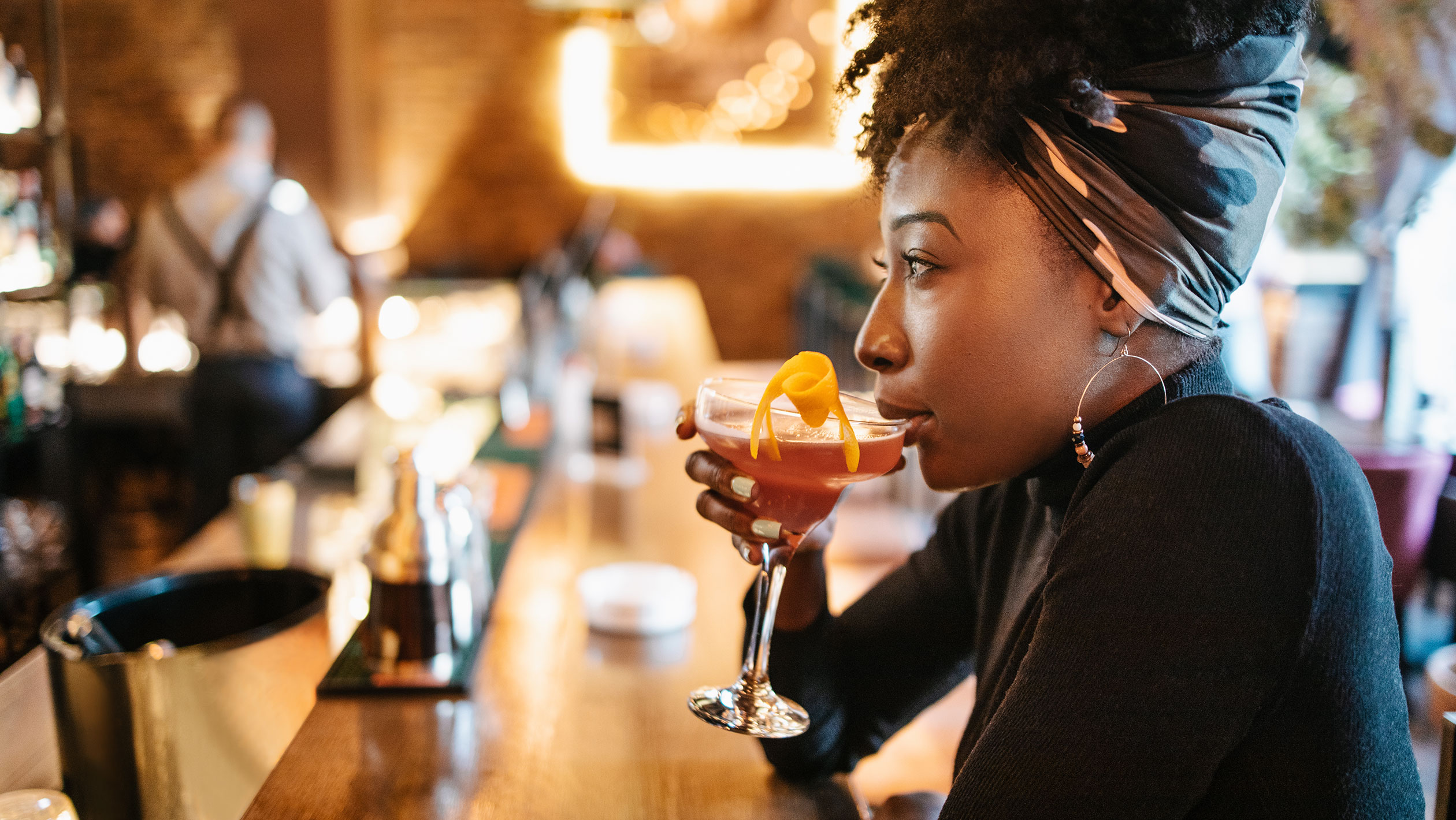 An African American woman sits at a bar and sips a cocktail
