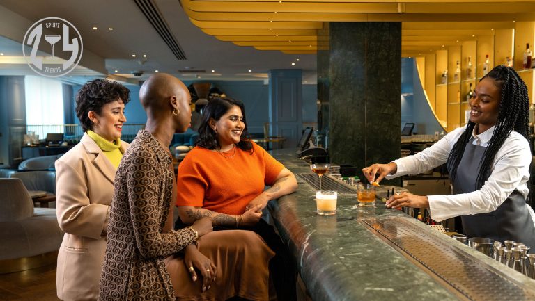 A group of women gather at a bar while a bartender serves their drinks. The SevenFifty Daily 2024 Trends logo stamp is in the upper left hand corner.