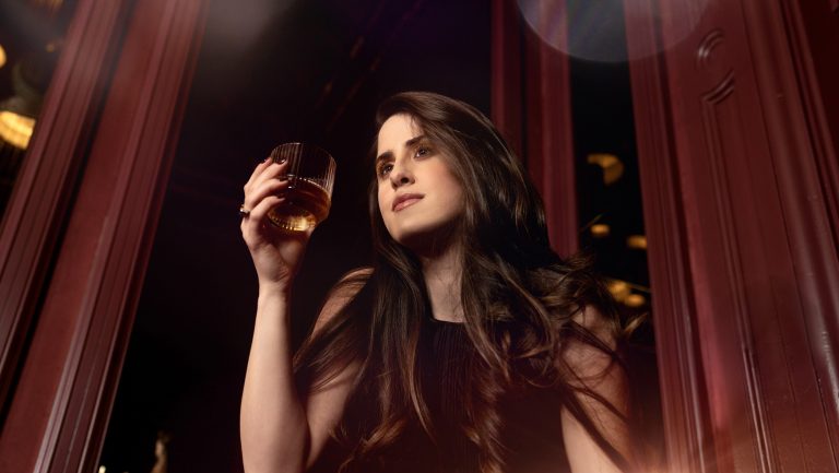 A woman poses with a glass of whisky in a warm toned, dimly lit setting