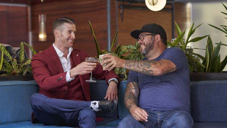 Josh Jacobs and Michael Bowen of Speakeasy Co toast their glasses while posing together on a couch