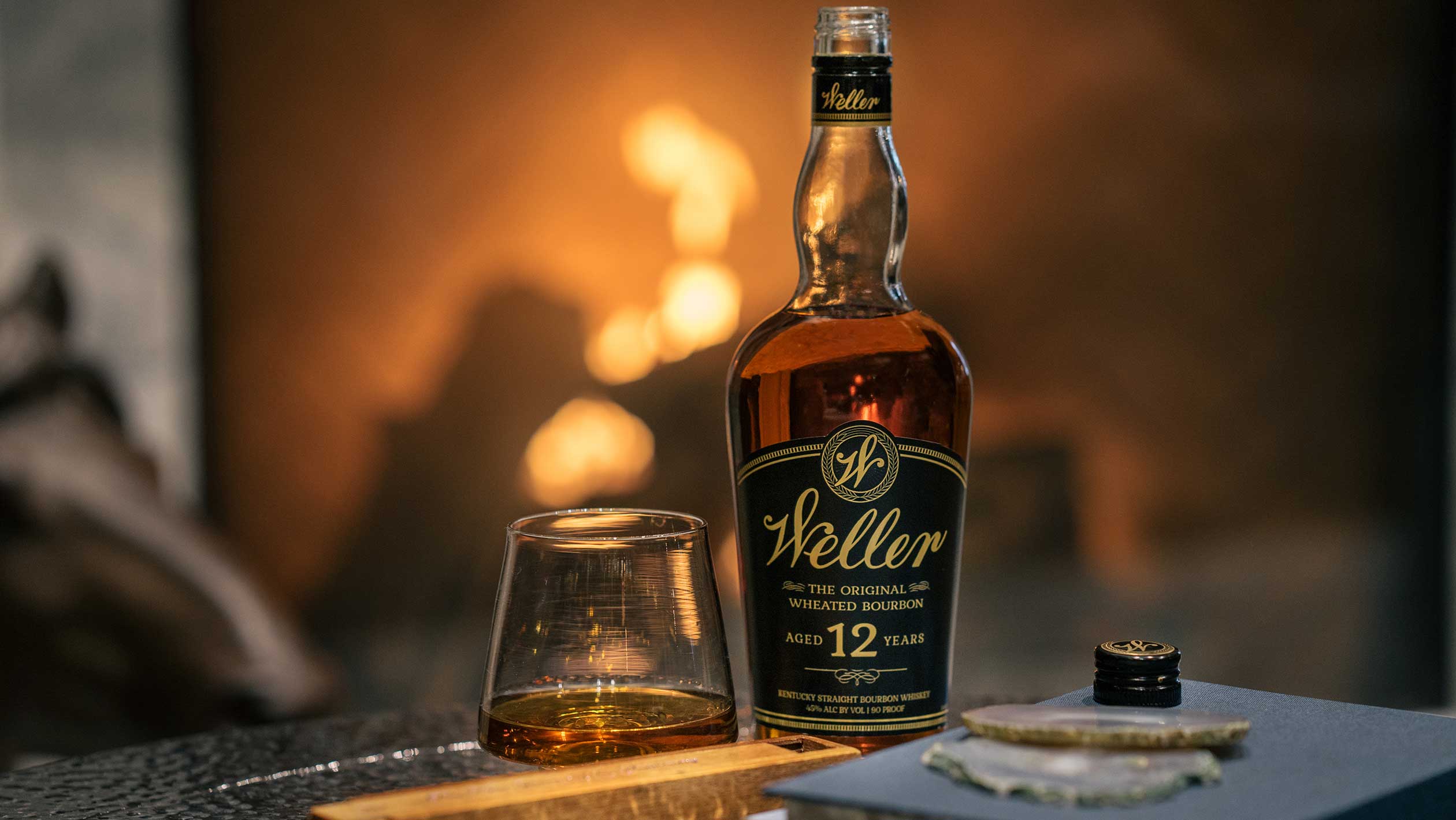 A bottle of Weller Original Wheated sits on a table with a roaring fireplace in the background