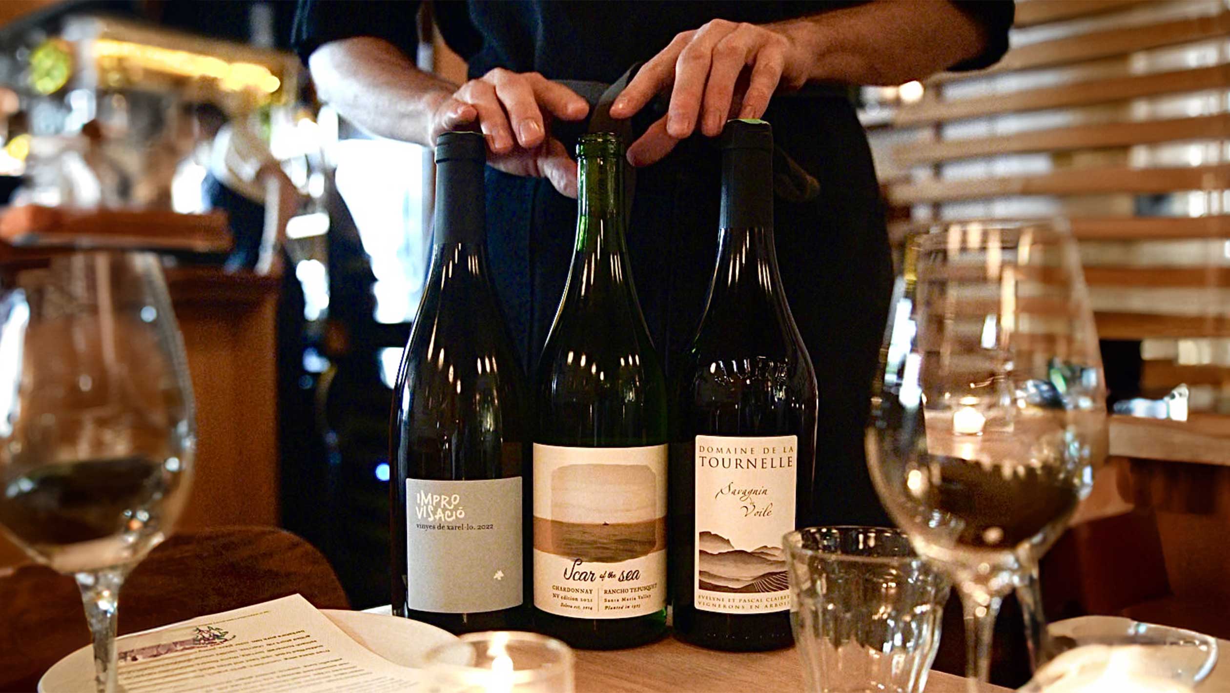 A person holds up three bottles of wine to the camera. From left to right: Enric Soler ‘Improvisacio’ 2022, Scar of the Sea ‘Rancho Tepusquet Solera’ NV, and Tournelle ‘Savagnin de Voile’ 2016.