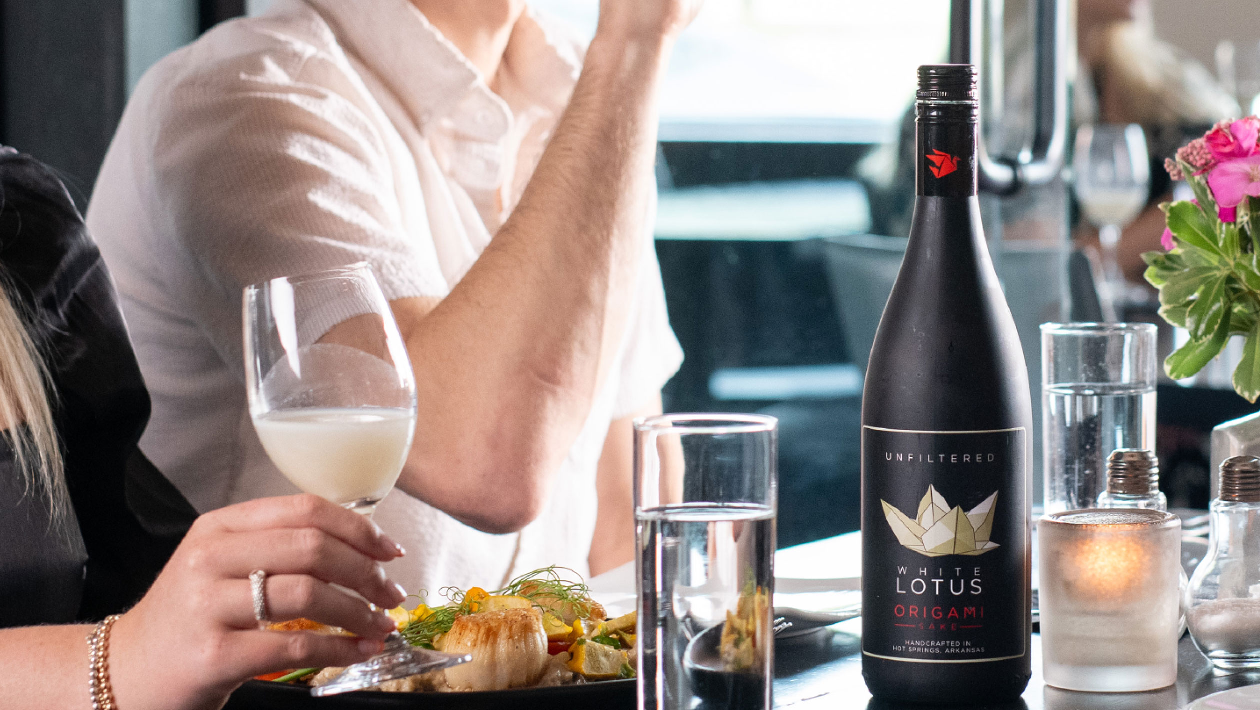 A bottle of Origami Sake is in the foreground of a table set with food and glasses of sake