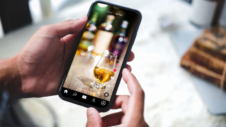 A pair of hands hold up a phone to show a TikTok feed with an alcohol ad playing