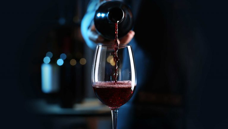 Red wine is poured into a wine glass in the foreground against a dark background