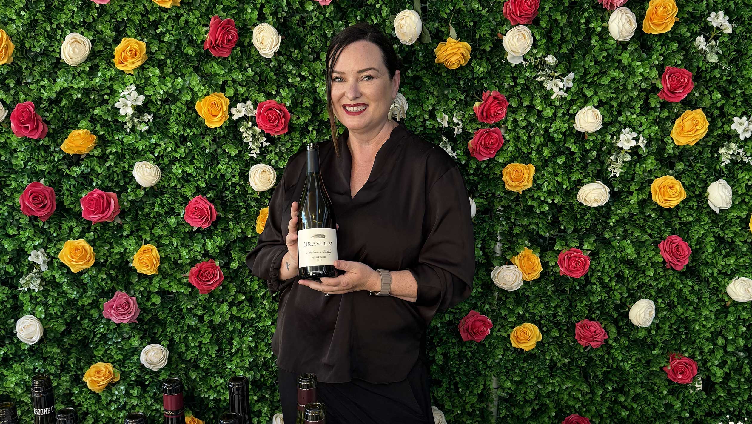 A picture of Melissa Smith posing with a wine bottle in front of a blooming hedge