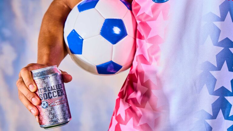 A photo of a soccer player holding a blue and white soccer ball and the special-edition beer "It's Called Soccer."