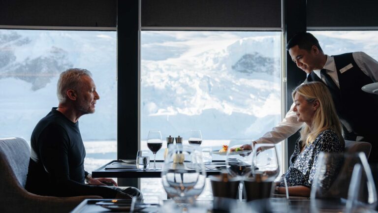 A photo of a man and a woman dining on a cruise ship, with wine on the table and glaciers in the background.